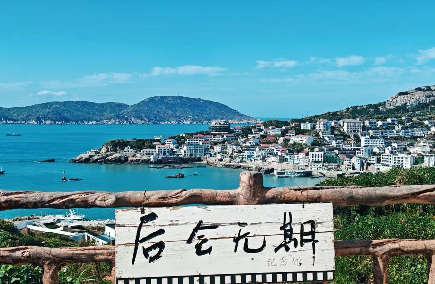 Dongji Island,Zhoushan,Chinatour Coastal town perched on hills with blue harbor water, seen behind a rustic wooden railing and a white sign with Chinese characters in the foreground.