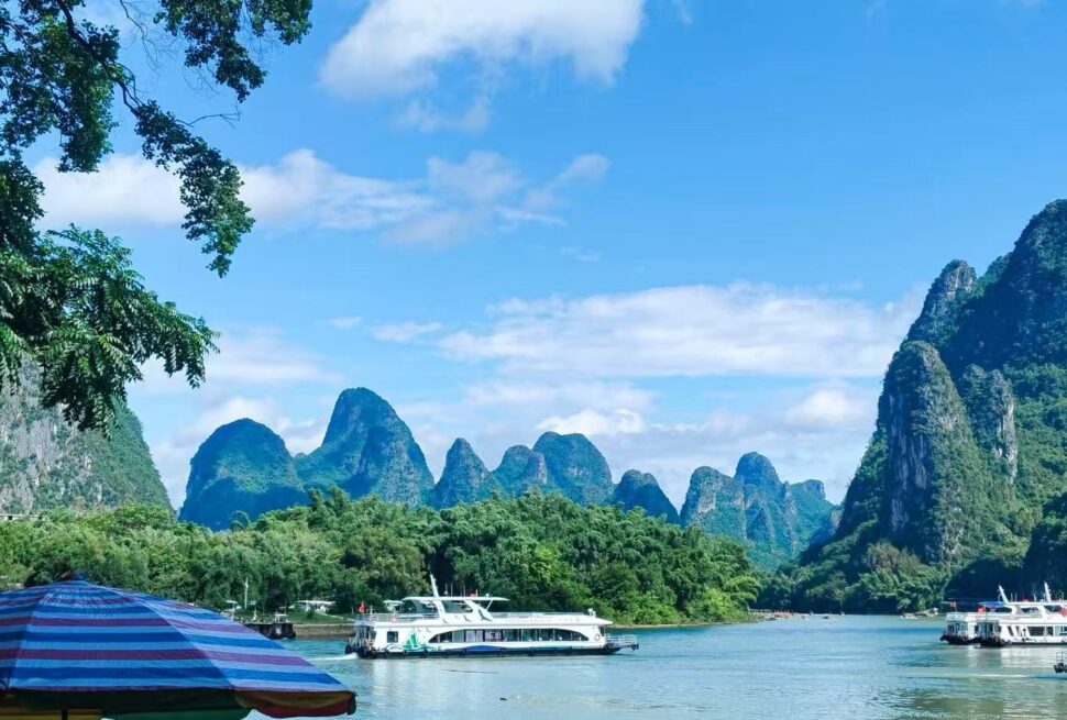 Scenic river lined with lush green trees and towering limestone karst hills under a blue sky; boats docked along the waterfront and a striped umbrella in the foreground.Xingping Ancient Town,Yangshuo,Guangxi,Chinatravel