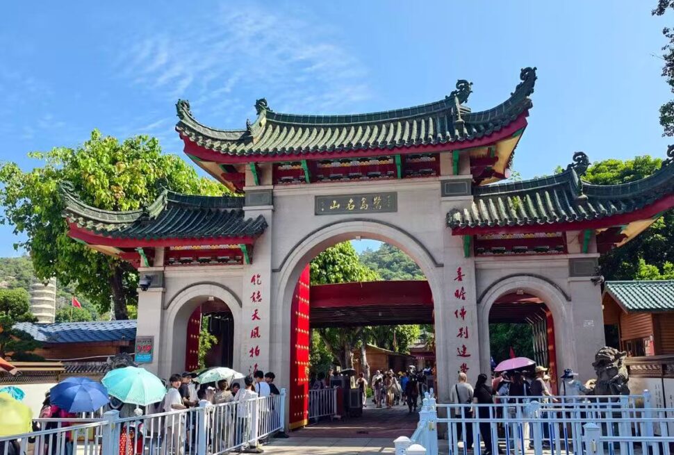 Nanputuo Temple,Xiamen,Chinatour Traditional Chinese temple entrance with curved roofs and red columns; people with umbrellas queue to enter on a sunny day.