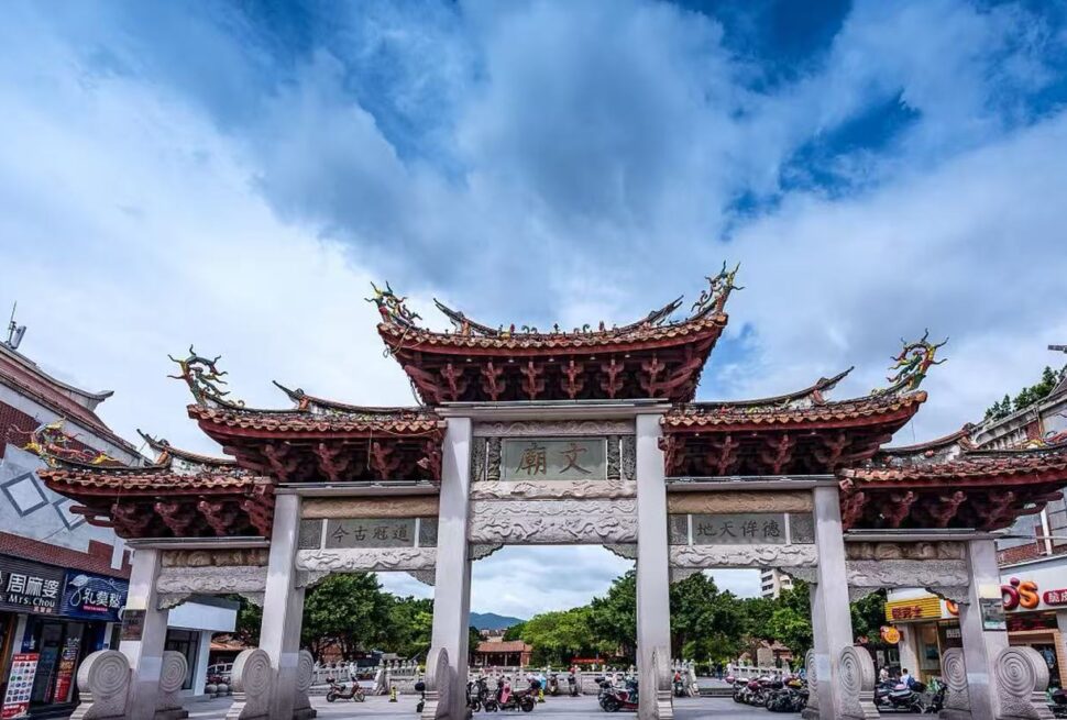 Stone ceremonial gate with ornate red-tiled Chinese roof in a busy street, shops on either side and scooters parked nearby.