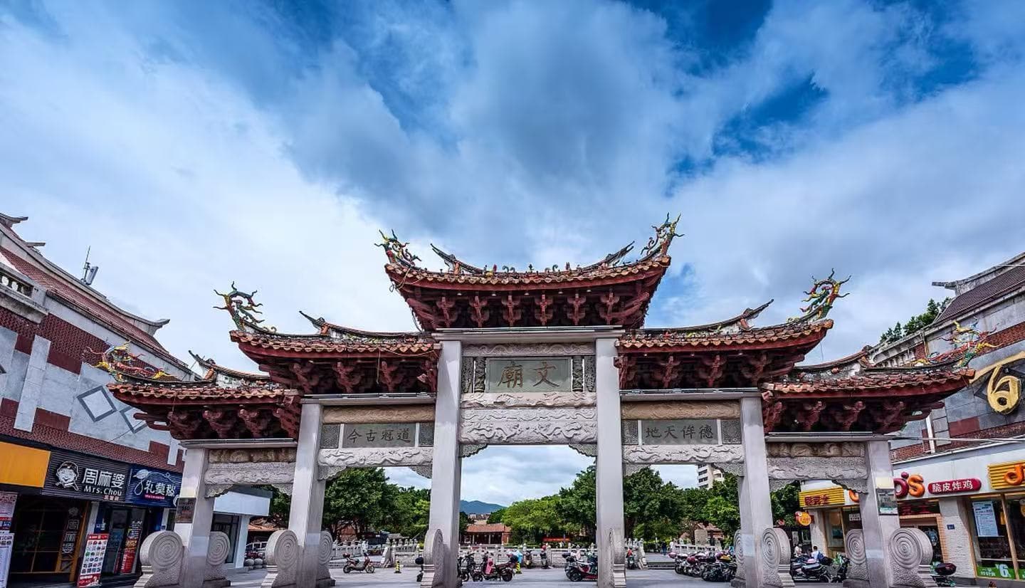 Stone ceremonial gate with ornate red-tiled Chinese roof in a busy street, shops on either side and scooters parked nearby.