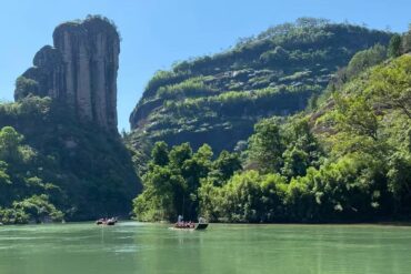 Wuyi Mountain UNESCO Site,Nanping,Chinatour Narrow river through a lush canyon with a tall rock pillar on the left and small boats on the water.