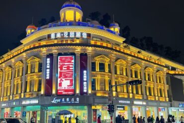 Nighttime view of a grand, neoclassical-style building illuminated in warm yellow lights, with a large red vertical billboard and people crossing the street at a busy intersection.