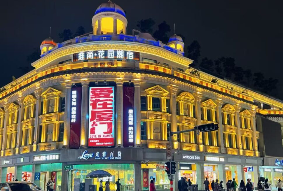 Nighttime view of a grand, neoclassical-style building illuminated in warm yellow lights, with a large red vertical billboard and people crossing the street at a busy intersection.