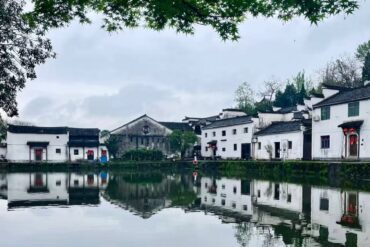Zhuge Bagua Village,Jinhua,Chinatour White-walled village by a calm lake with mirrored reflections, gray sky, and overhanging green branches framing the scene.
