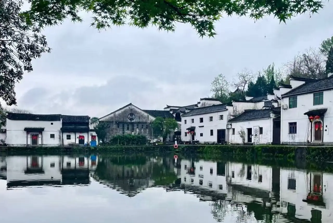 Zhuge Bagua Village,Jinhua,Chinatour White-walled village by a calm lake with mirrored reflections, gray sky, and overhanging green branches framing the scene.