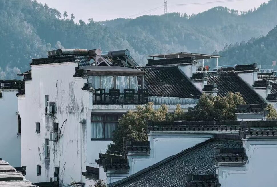 White-walled houses with dark tiled roofs clustering on a hillside, with trees in front and forested mountains behind them.