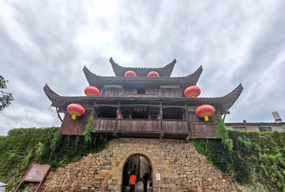 Traditional wooden gate tower atop a stone archway, adorned with red lanterns and creeping ivy against a cloudy sky treated as an historic entrance.Hepingi Ancient Town,Nanping,Fujian,Chinatravel