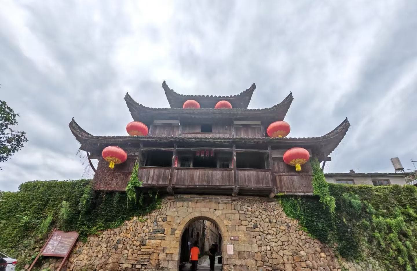 Traditional wooden gate tower atop a stone archway, adorned with red lanterns and creeping ivy against a cloudy sky treated as an historic entrance.Hepingi Ancient Town,Nanping,Fujian,Chinatravel