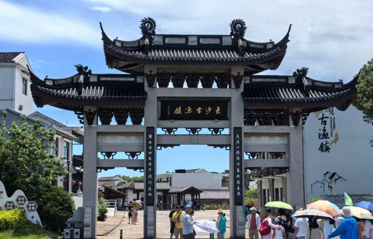 Dongsha Ancient Town,Zhoushan,Chinatour Traditional Chinese arch gate with ornate curved roofs and a dark plaque, visitors entering a cultural town under a blue sky.
