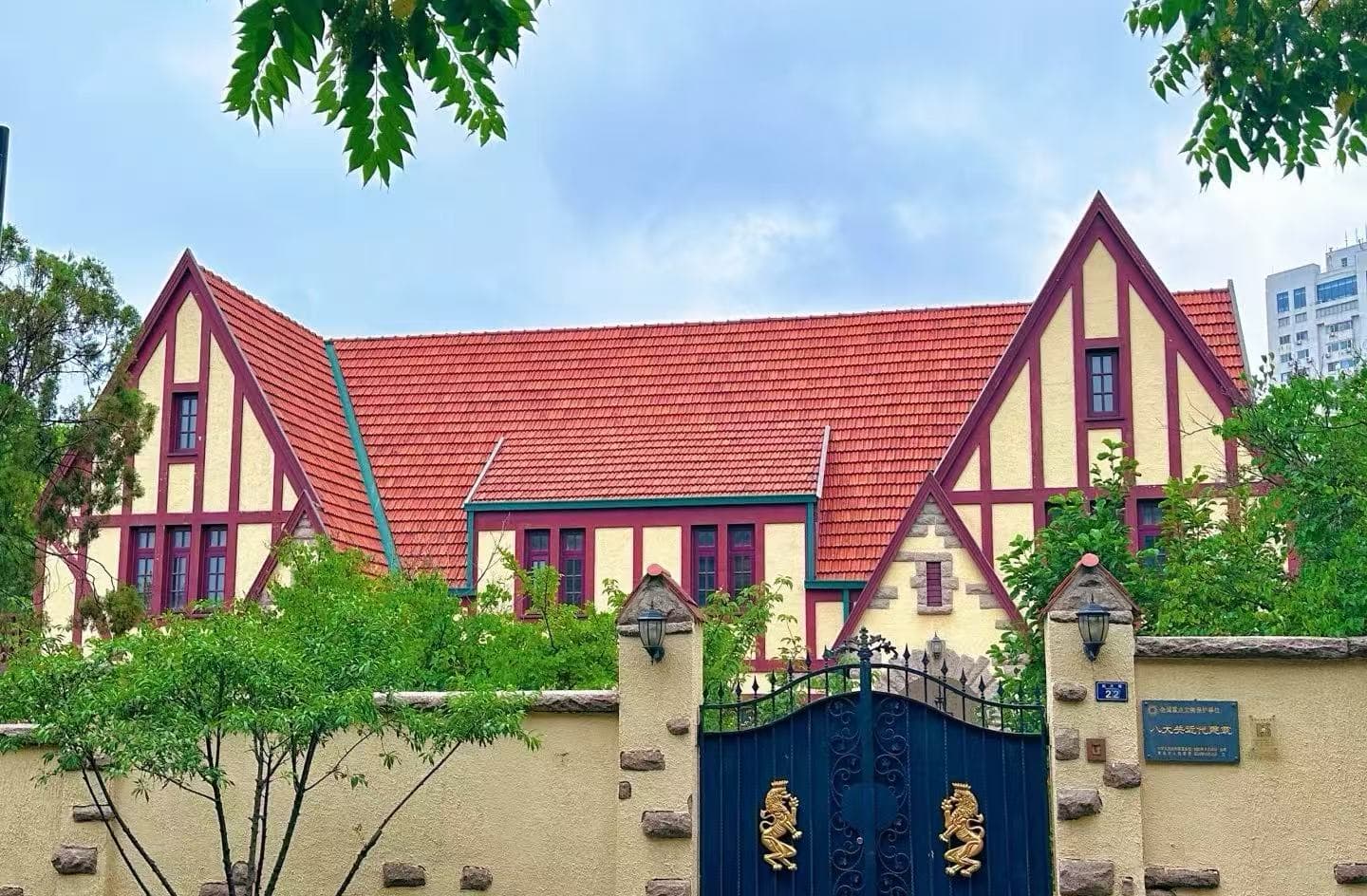 Badaguan Scenic Area,Qingdao,Chinatour Colorful European-style house with yellow walls, red trim and steep red-tiled roof behind a stone fence and blue gate with dragon symbols, greenery in front.
