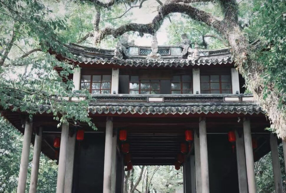 Fenghua Xikou Ancient Town,Ningbo,Chinatour Traditional Chinese pavilion entrance with red lanterns under a tiled roof, surrounded by dense greenery and trees.