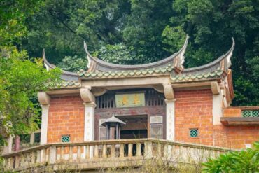 Traditional Chinese temple gate with curved tiled roof, red brick walls, and stone balustrade amid lush greenery