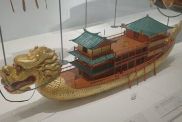 Quanzhou Maritime Museum,Quanzhou,Chinatour Ornate gold dragon-headed boat model with multiple teal-roofed pavilions on its back, displayed in a glass case.