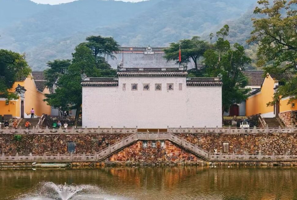 Tiantong Temple,Ningbo,Chinatour Temple complex by a pond with a white central wall, orange side buildings, and green trees against a mountainous backdrop.