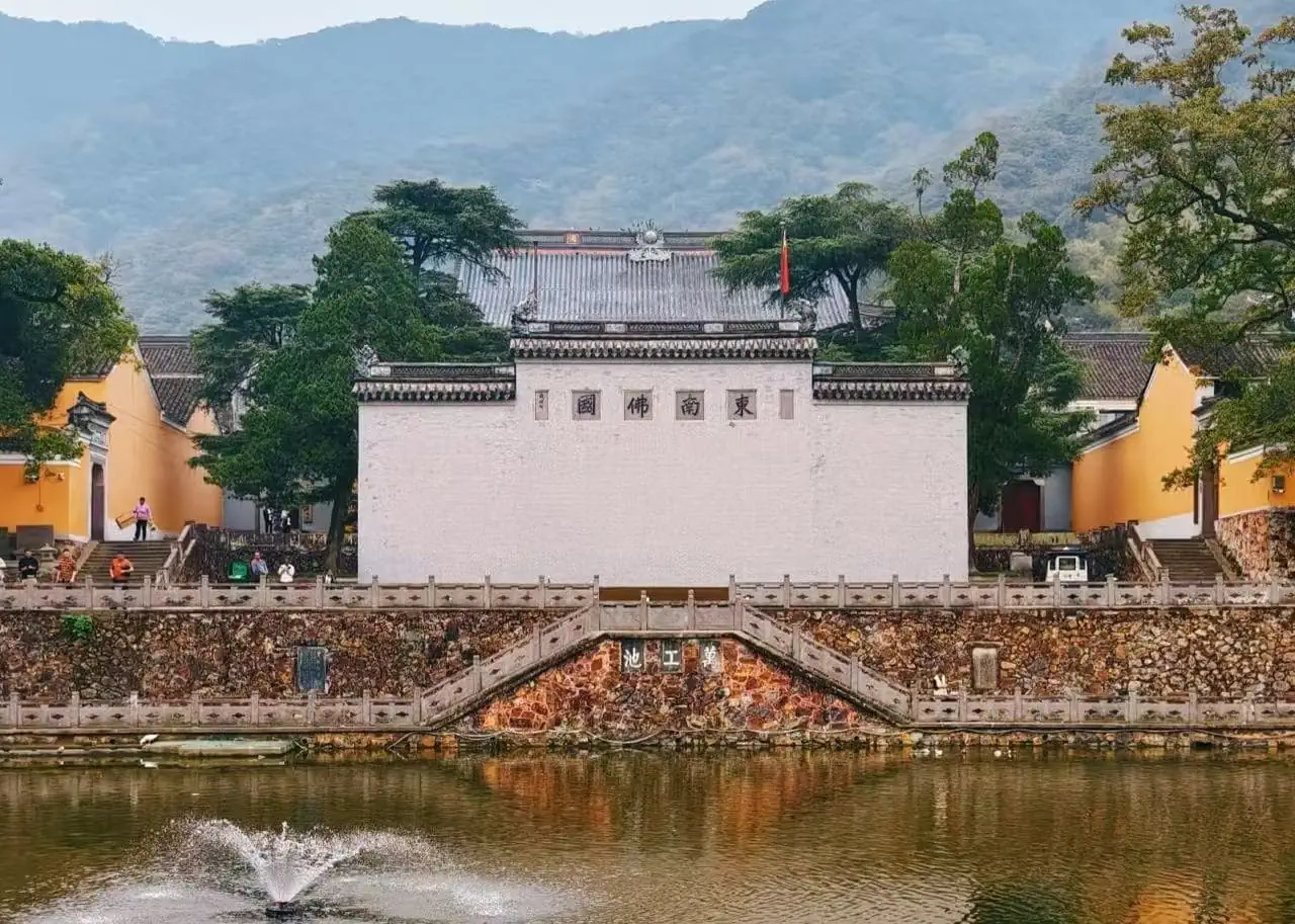 Tiantong Temple,Ningbo,Chinatour Temple complex by a pond with a white central wall, orange side buildings, and green trees against a mountainous backdrop.