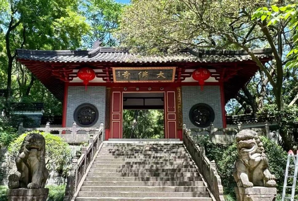 Great Buddha Temple ,Shaoxing,Chinatour Entrance to a traditional Chinese temple, red wooden gates atop stone steps, flanked by two stone guardian lion statues and hanging red lanterns in a lush garden setting.