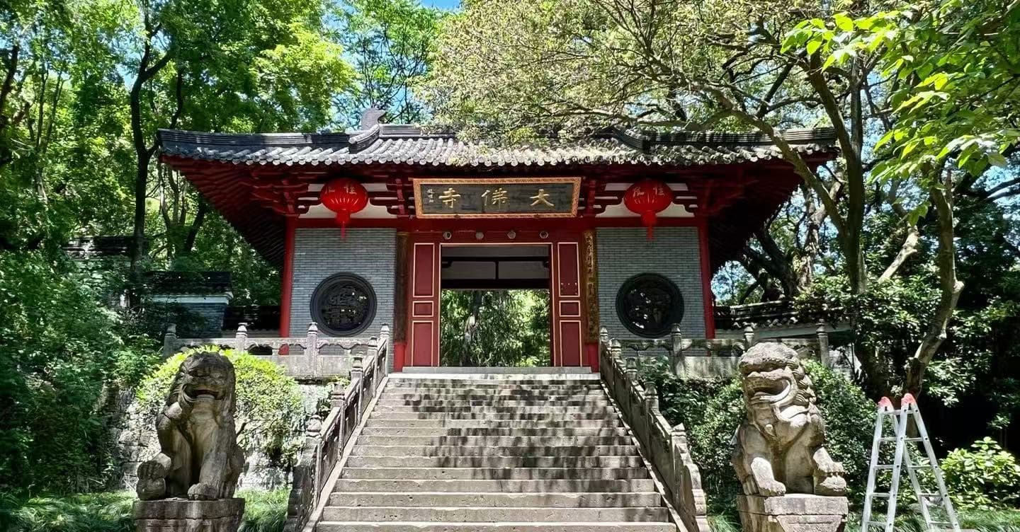 Entrance to a traditional Chinese temple, red wooden gates atop stone steps, flanked by two stone guardian lion statues and hanging red lanterns in a lush garden setting.