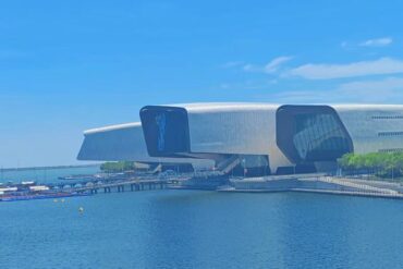 National Maritime Museum,Tianjin,Chinatour Futuristic silver cultural building with curved, wing-like structures along a calm blue waterfront and a wooden boardwalk with boats nearby.