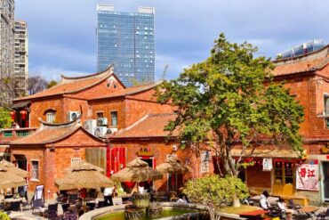 Traditional red-brick Chinese courtyard with thatched umbrellas, outdoor seating, and a stone fountain; modern high-rise in the background with blue sky behind.