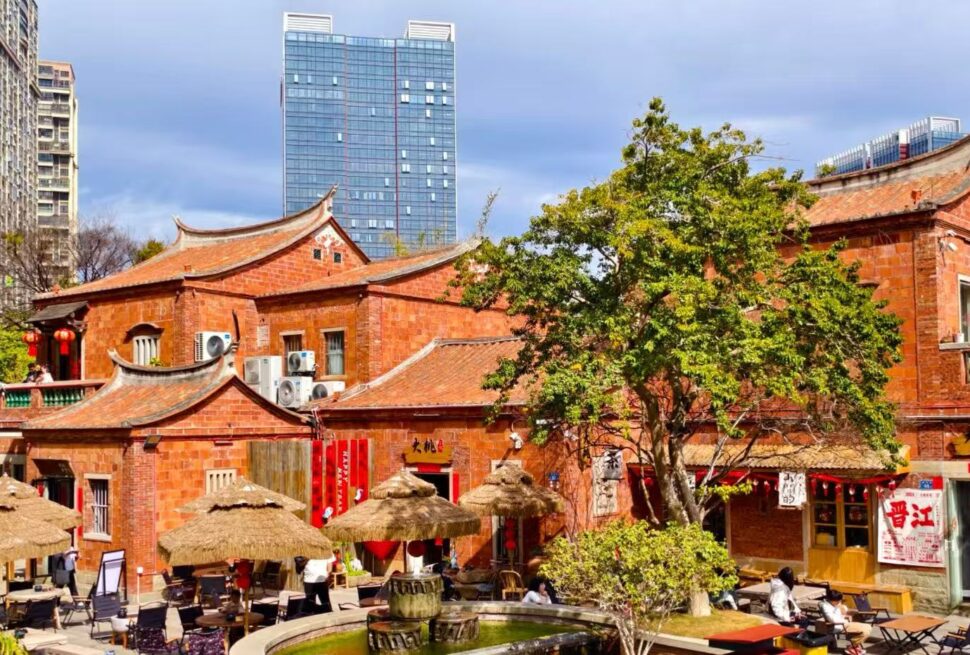 Traditional red-brick Chinese courtyard with thatched umbrellas, outdoor seating, and a stone fountain; modern high-rise in the background with blue sky behind.