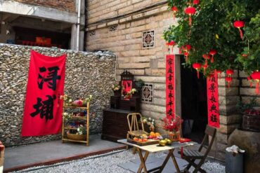 Red vertical Chinese New Year couplets hang by a stone wall beside a small wooden altar with flowers and fruit offerings on a folding table outside a doorway.