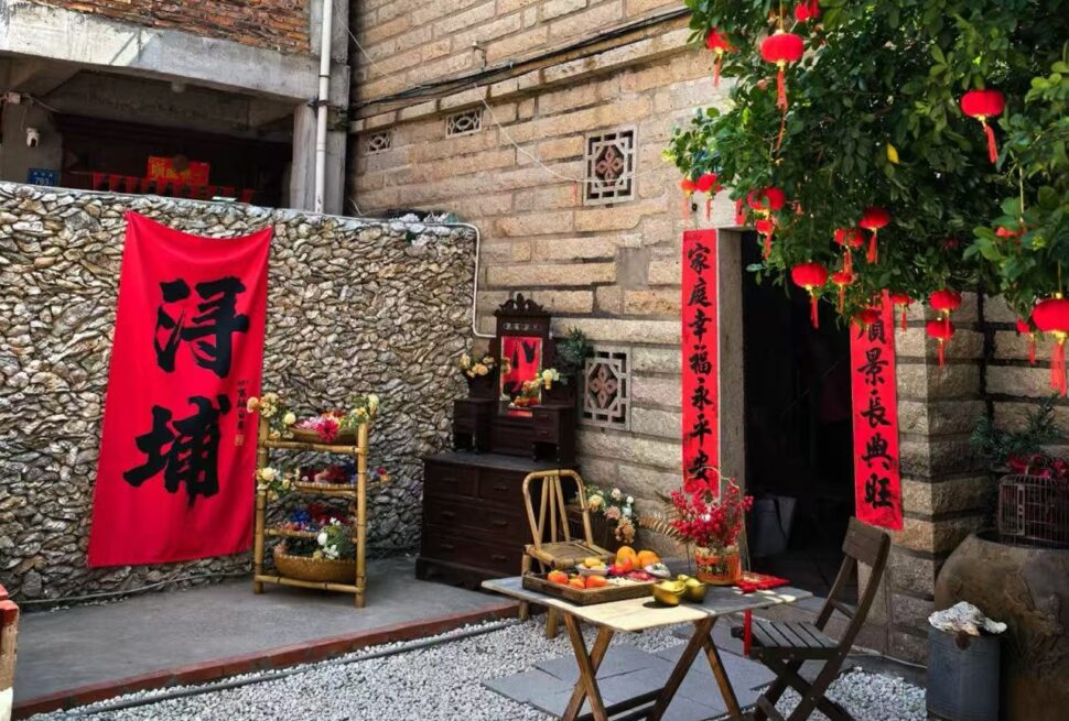 Xunpu Village ,Quanzhou,Chinatour Red vertical Chinese New Year couplets hang by a stone wall beside a small wooden altar with flowers and fruit offerings on a folding table outside a doorway.