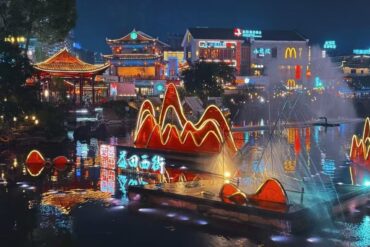 Nighttime river scene with glowing red neon mountain sculptures on the water and traditional Chinese architecture in the background.Yangshuo West Street,Yangshuo,Guangxi,Chinatravel