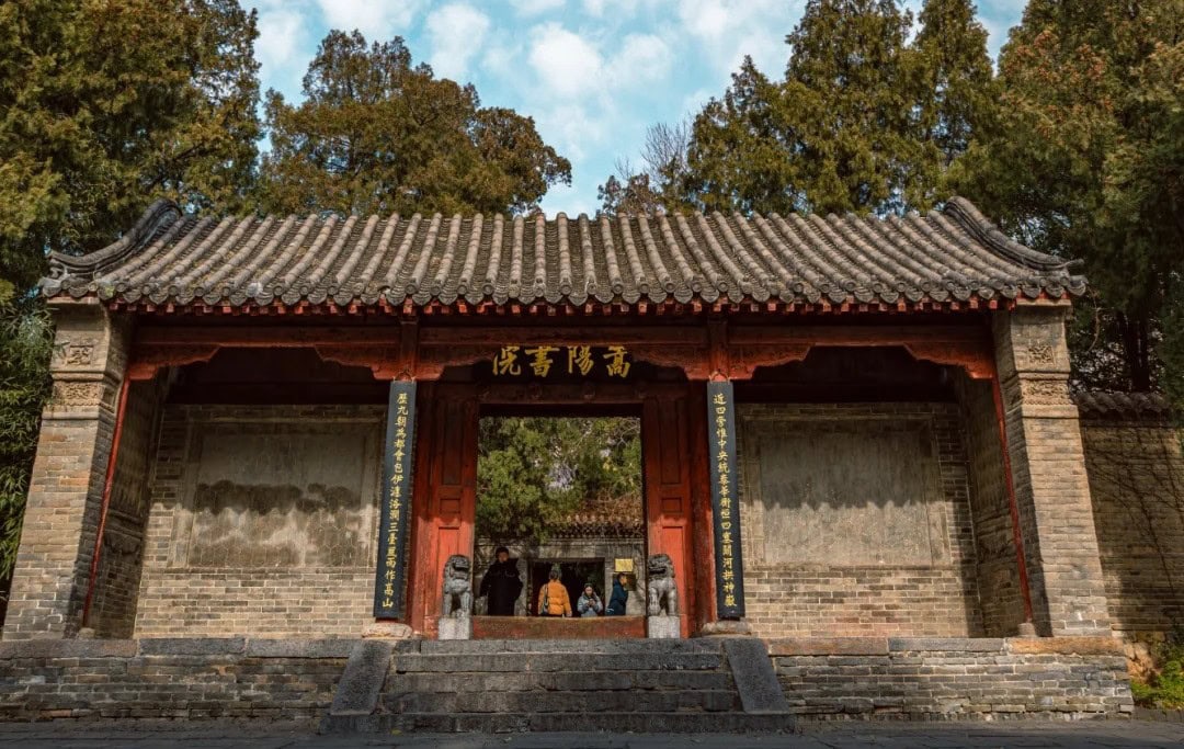 Traditional Chinese temple gate with a tiled roof, red pillars, gold inscription, guardian lions, and visitors standing under the doorway.Songyang Academy,Shaolin & Songshan Area,Henan,Chinatravel