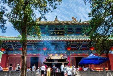 Ornate Chinese temple facade with a teal roof, red walls, hanging red lanterns, and worshippers at the front altar, smoke rising from incense.Songshan (Song Mountains) Scenic Area,Shaolin & Songshan Area,Henan,Chinatravel