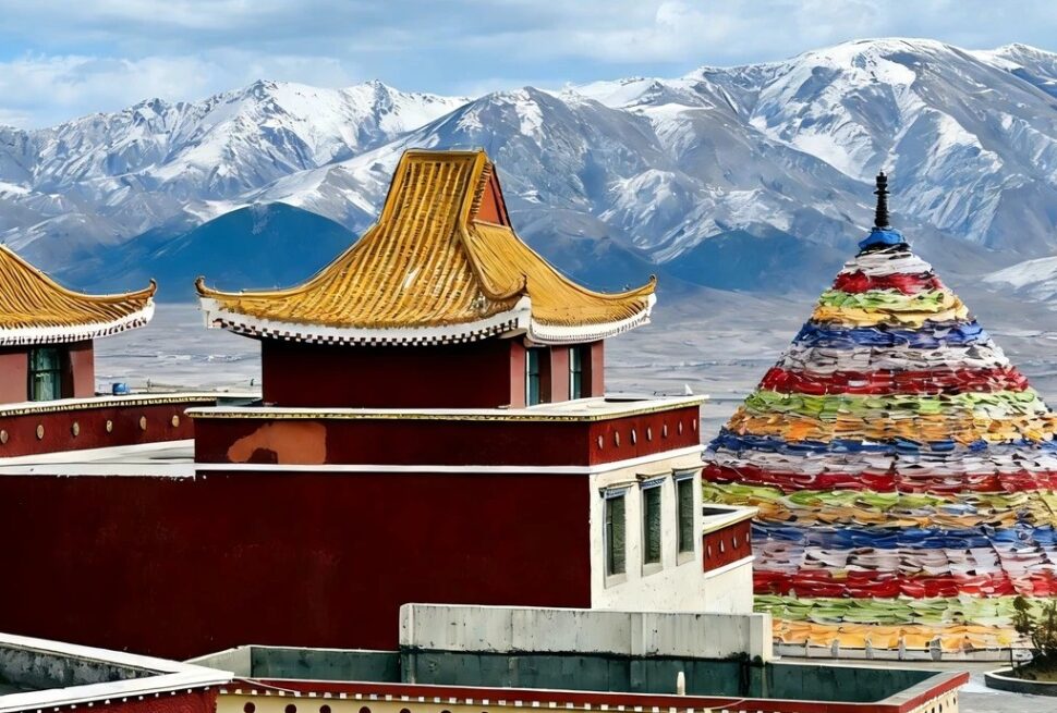 Red and gold-trimmed temple buildings with tiered roofs set against snow-capped mountains and a colorful prayer-stone cone nearby, Tibet.Riyue Mountain Pass,Xining,Qinghai