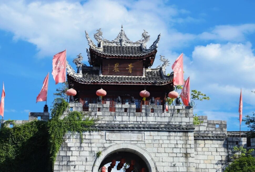 Traditional Chinese temple gate atop a stone archway with red lanterns and pink banners against a blue sky.Qingyan Ancient Town,Guiyang,Guizhou