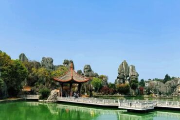 A traditional Chinese-style pavilion with red roof on a wooden jetty over a calm green pond, surrounded by rocky formations and trees.Kunming Stone Forest,Kunming,Yunnan ,Chinatravel