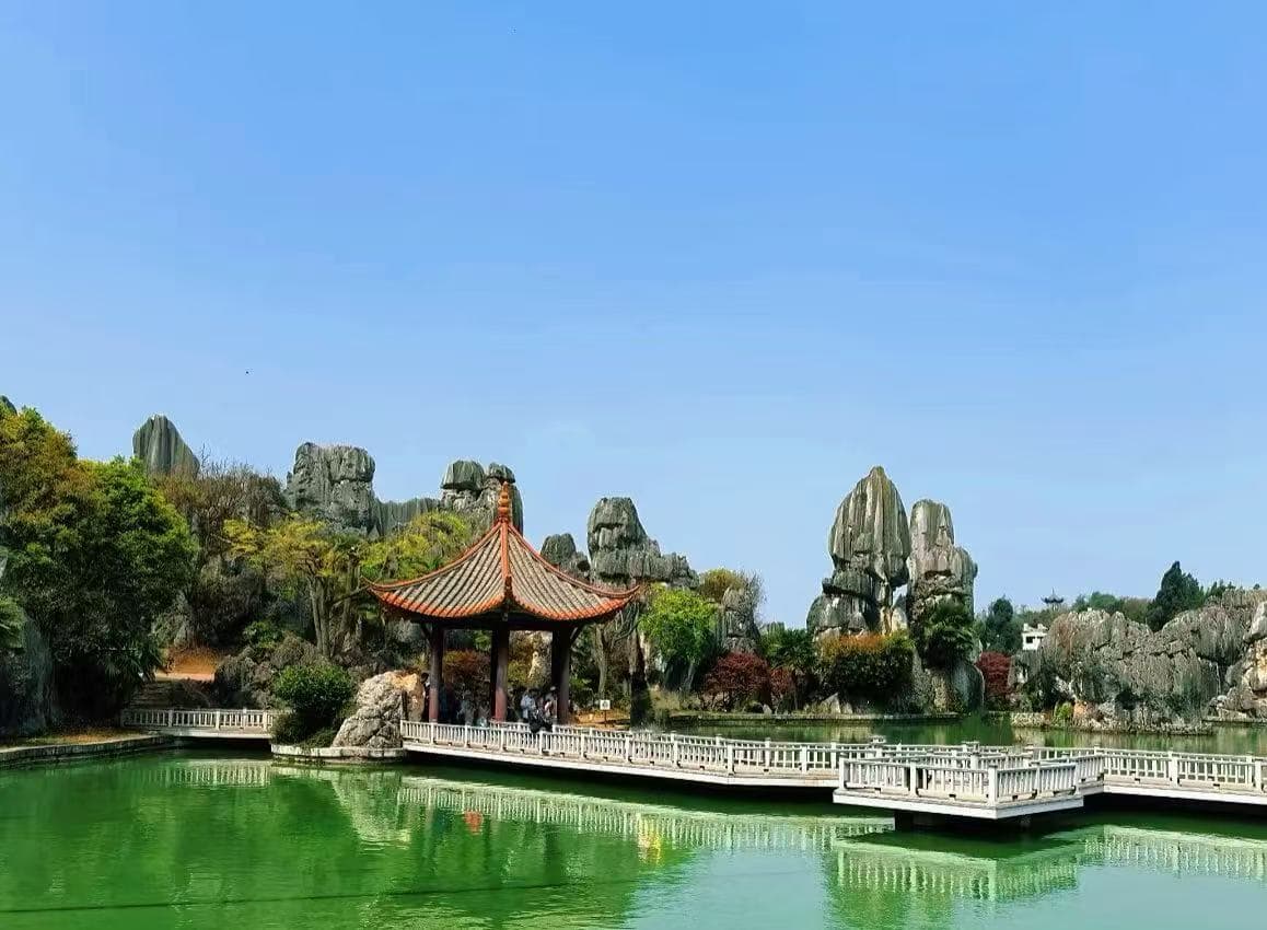 A traditional Chinese-style pavilion with red roof on a wooden jetty over a calm green pond, surrounded by rocky formations and trees.Kunming Stone Forest,Kunming,Yunnan ,Chinatravel