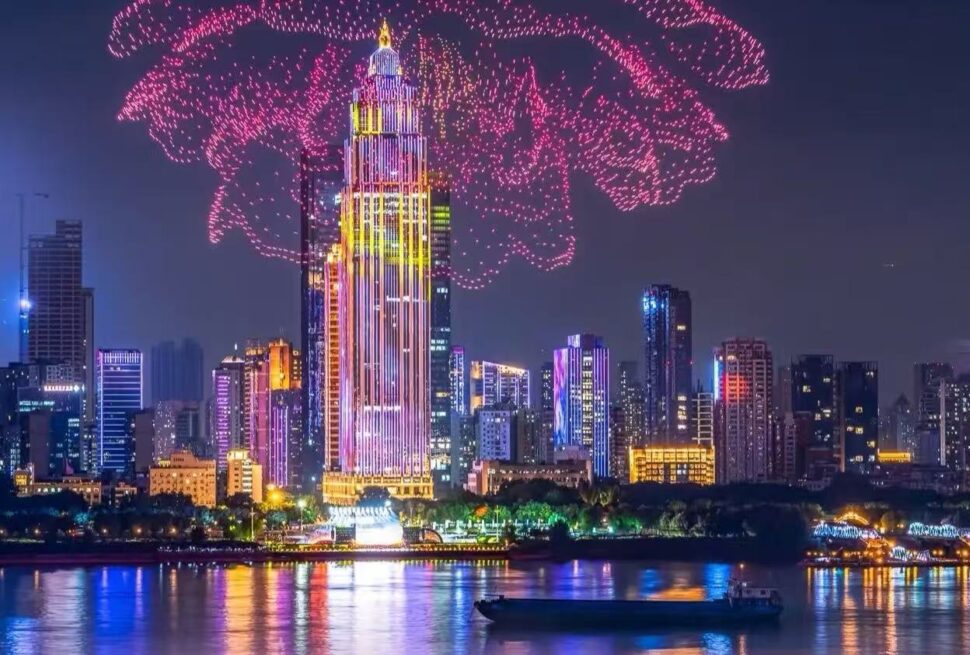 Nighttime city skyline with a tall illuminated central skyscraper, pink fireworks overhead, and reflections on the river with a boat in the foreground.Yangtze River Cruise,Wuhan,Hubei ,Chinatravel