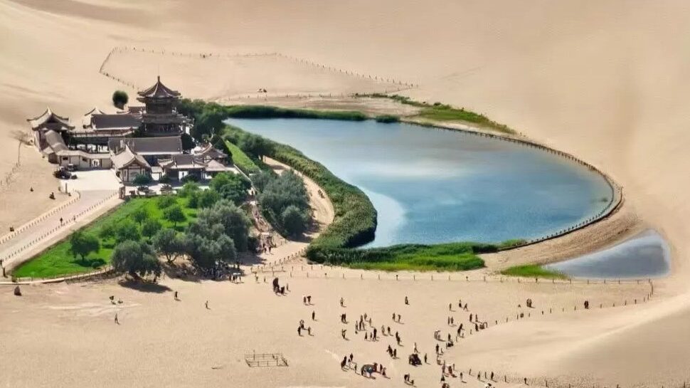 Desert oasis with a circular blue lake surrounded by green lawns and a traditional Chinese temple complex nestled in sand dunes.Crescent Lake & Mingsha Mountain,Dunhuang,Gansu