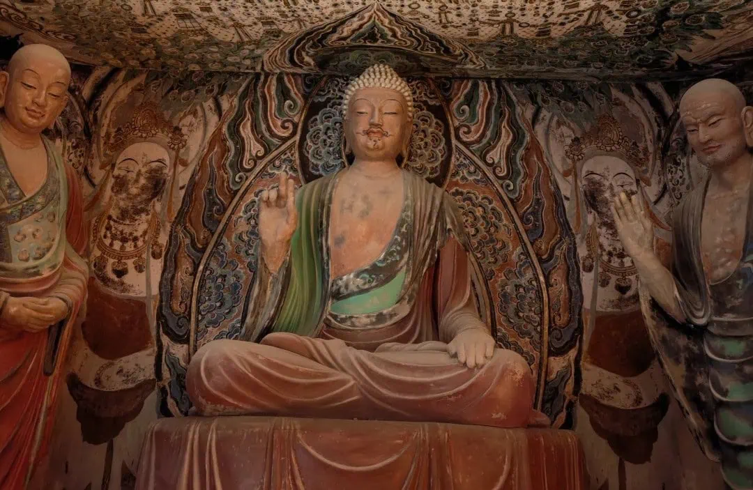Central seated Buddha statue in a richly painted cave temple, with two standing figures on either side.Dunhuang Museum,Dunhuang,Gansu