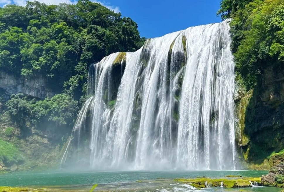 Waterfall cascades down a rocky cliff into a turquoise pool, surrounded by lush green forest on a sunny day.Huangguoshu Waterfall,Guiyang,Guizhou