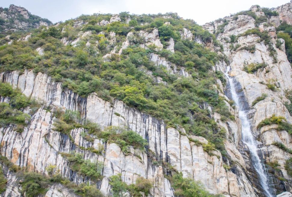 Steep rocky mountainside with striped vertical rock faces, green shrubs, and a narrow waterfall on the right side.Songshan Luya Waterfall,Shaolin & Songshan Area,Henan,Chinatravel