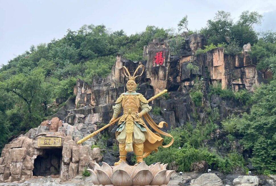 Golden armored warrior statue holding a long staff, standing on a lotus pedestal at a rocky hillside with greenery and a cave entrance behind him.Jilin Beishan Park,Chinatravel