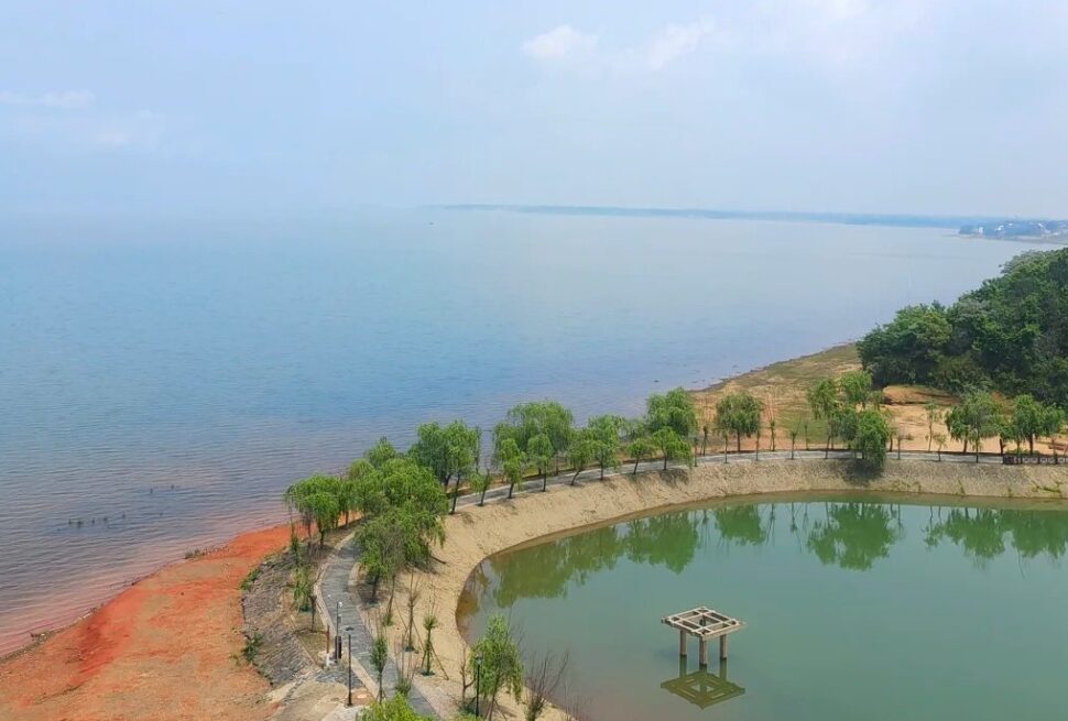 A curved shoreline with a green pond, lined by trees and a walking path, meeting a calm sea in the distance.Poyang Lake National Wetland Park,Shangrao,Jiangxi,Chinatravel