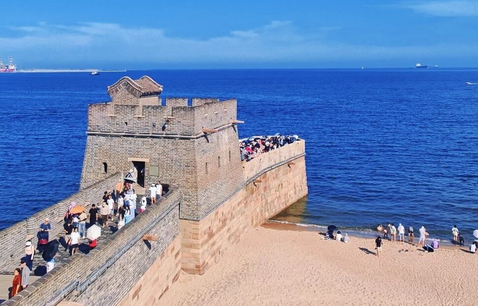 Stone coastal fortress with a walkway filled with visitors along the wall, overlooking a calm blue sea on a sunny day.Shanhaiguan,Qinhuangdao,Hebei,Chinatravel