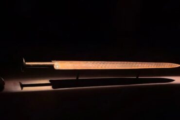 Ornate dagger with a metal hilt and long slim blade, placed above a brown sheath on a dark surface.Hubei Provincial Museum,Wuhan,Hubei ,Chinatravel