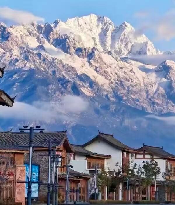 Snow-capped mountains tower over a street of traditional wooden buildings, blue sky and light clouds above.Jade Dragon Snow Mountain,Lijiang,Yunnan,Chinatravel