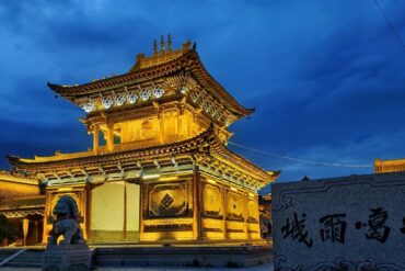Illuminated ancient Chinese temple gate with ornate, multi-tiered roof at dusk, flanked by stone lion statues.Donggar Ancient City,Xining,Qinghai