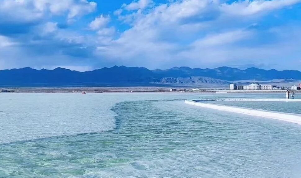 Turquoise shallow water with a curved white boardwalk leading to distant buildings and blue mountain range under a partly cloudy sky,Chaka Salt Lake,Xining,Qinghai