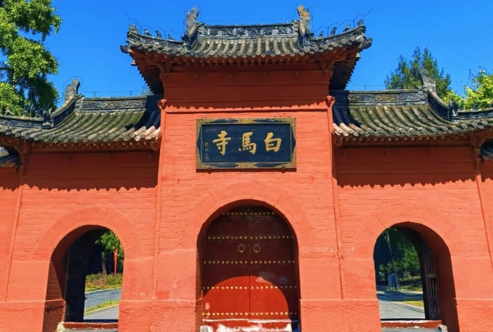 Red temple gate with curved gray-tiled roof, orange-red walls, and a central arched door beneath a black plaque with gold Chinese characters.
