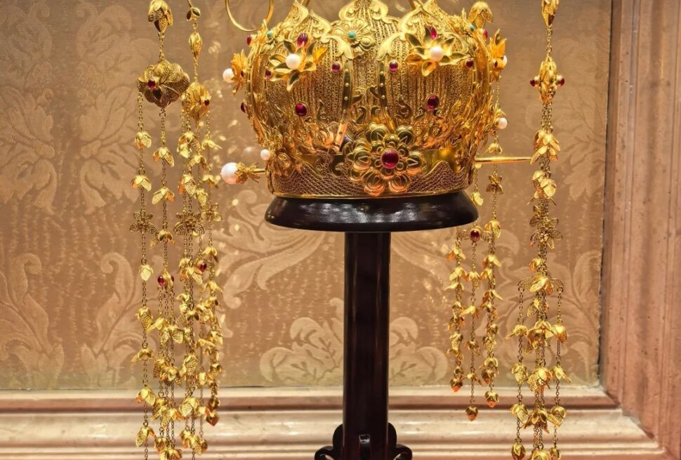 Ornate gold crown encrusted with red gems and pearls, displayed on a black pedestal with hanging gold chains around it.Guiyang White House,Guiyang,Guizhou