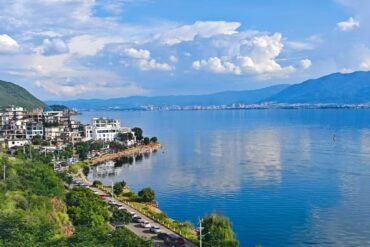 Lakeside town with white buildings along a rocky shore, green hills, and a calm blue lake with distant mountains under a cloudy sky.Erhai Lake,Dali,Yunnan,Chinatravel