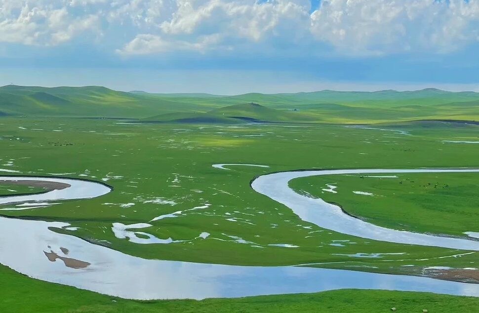 Mergel Gol River,Hulunbuir,Chinatour Wide green plain with a winding river curving through fields under a cloudy sky.